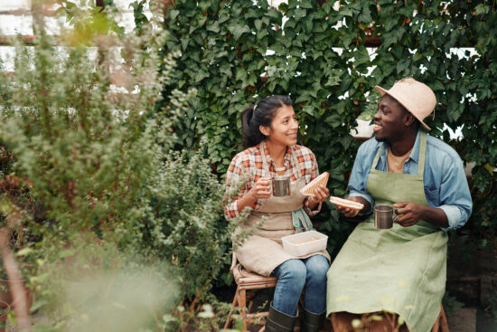 Two young ethnically diverse co-workers wearing aprons enjoying having lunch together in modern greenhouse