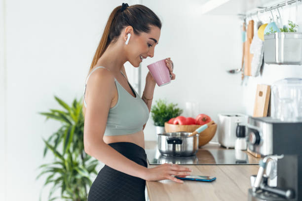 A sporty woman listening to music with her smartphone while drinking coffee in the kitchen.