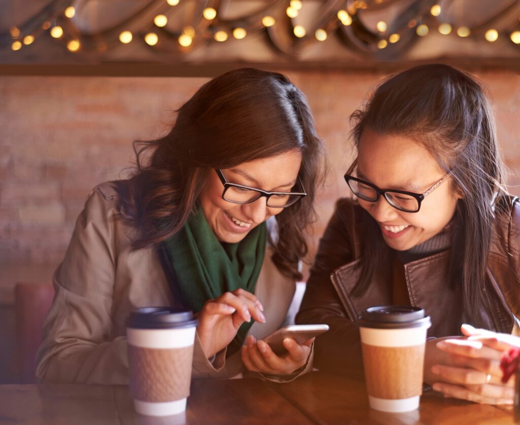 two gen z girls smiling and looking at something on a cellphone while drinking coffee