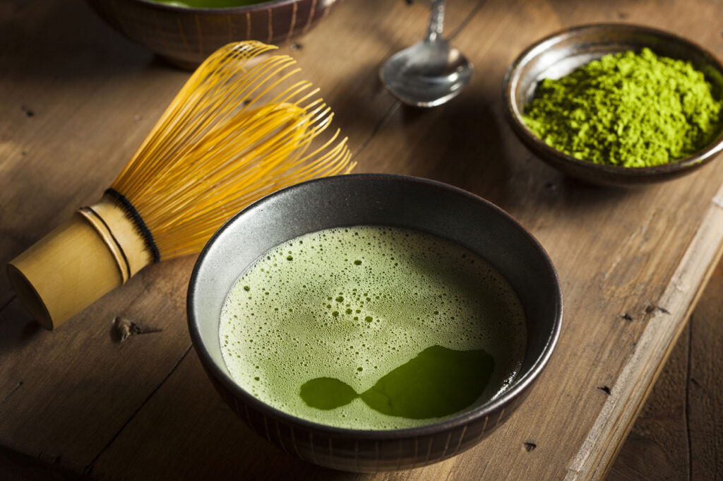 Matcha powder and matcha liquid in bowls, with a matcha whisk on a table