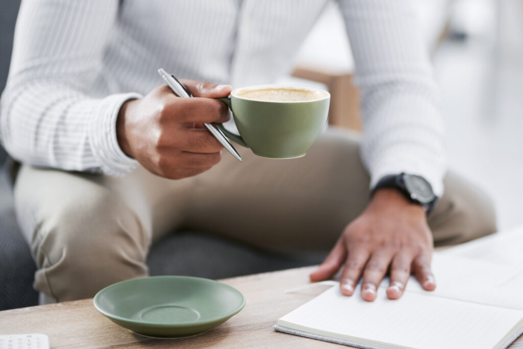 Closeup shot of an unrecognisable businessman drinking coffee while writing notes in an office.