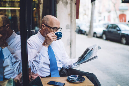 Elderly short haired man in white shirt sitting and reading newspaper while drinking coffee from cup on city street near glass and road